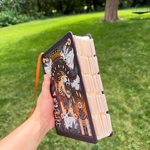 May include: A black Holy Bible with an intricate graphic illustration featuring a roaring lion, a crown, and floral motifs in orange, white, and gold. The book includes a bright orange ribbon bookmark and white index tabs along the side edge.