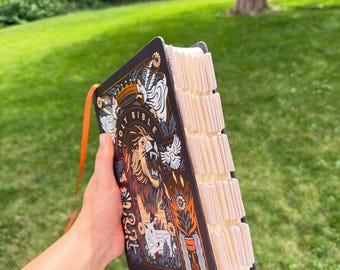 May include: A black Holy Bible with an intricate graphic illustration featuring a roaring lion, a crown, and floral motifs in orange, white, and gold. The book includes a bright orange ribbon bookmark and white index tabs along the side edge.