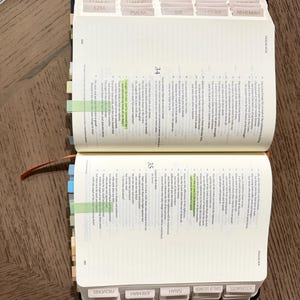 May include: An open Bible with white pages, black printed text, and numerous light-colored adhesive index tabs along the edges. Two specific verses are highlighted in neon green. The book features a dark cover and an orange ribbon bookmark.