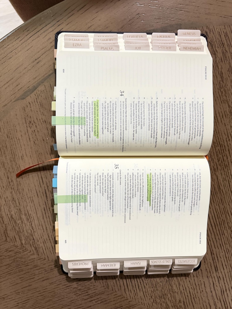 May include: An open Bible with white pages, black printed text, and numerous light-colored adhesive index tabs along the edges. Two specific verses are highlighted in neon green. The book features a dark cover and an orange ribbon bookmark.