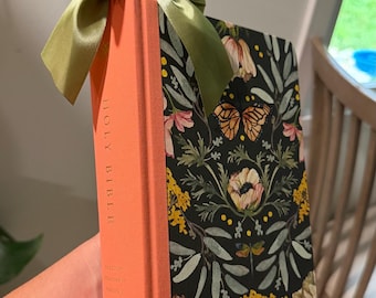 May include: A Holy Bible with a coral-colored spine and a black cover featuring a botanical pattern of flowers, leaves, and butterflies. A sage green satin ribbon is tied in a bow around the top of the book spine. The text on the spine reads Holy Bible, English Standard Version, and Crossway.