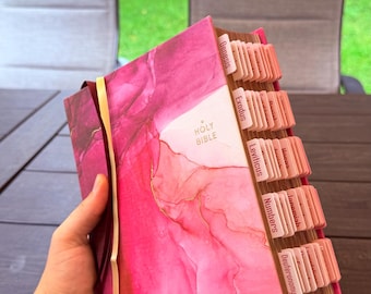 May include: A Holy Bible with a vibrant pink and magenta alcohol ink-style cover featuring gold accents. The book includes numerous light pink index tabs along the side, each labeled with the names of various books of the Bible, and a ribbon.