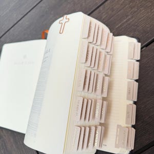 May include: A Bible with cream-colored pages featuring a series of organized, light-colored index tabs along the side. A small, metallic, rose gold-colored cross-shaped paperclip is attached to the top edge of the pages to mark a specific section.
