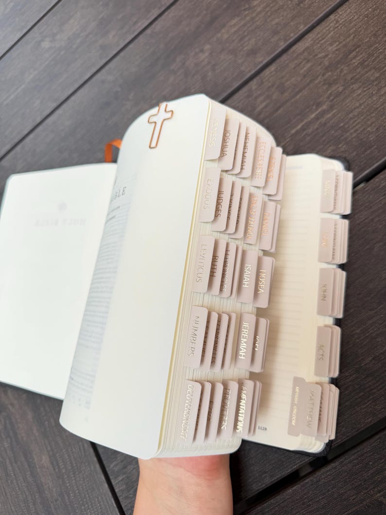 May include: A Bible with cream-colored pages featuring a series of organized, light-colored index tabs along the side. A small, metallic, rose gold-colored cross-shaped paperclip is attached to the top edge of the pages to mark a specific section.