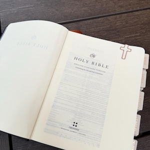 May include: An open Holy Bible, English Standard Version, featuring a rose gold cross-shaped paperclip on the top right page. The book includes light gray index tabs labeled Genesis, Exodus, Leviticus, Numbers, and Deuteronomy along the side.