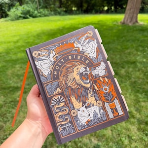 May include: A dark gray hardcover Holy Bible featuring a detailed graphic illustration of a roaring lion, a lamb, angels, and a sword. The cover design uses orange, white, and gray ink. The book includes a bright orange ribbon bookmark and tabs.