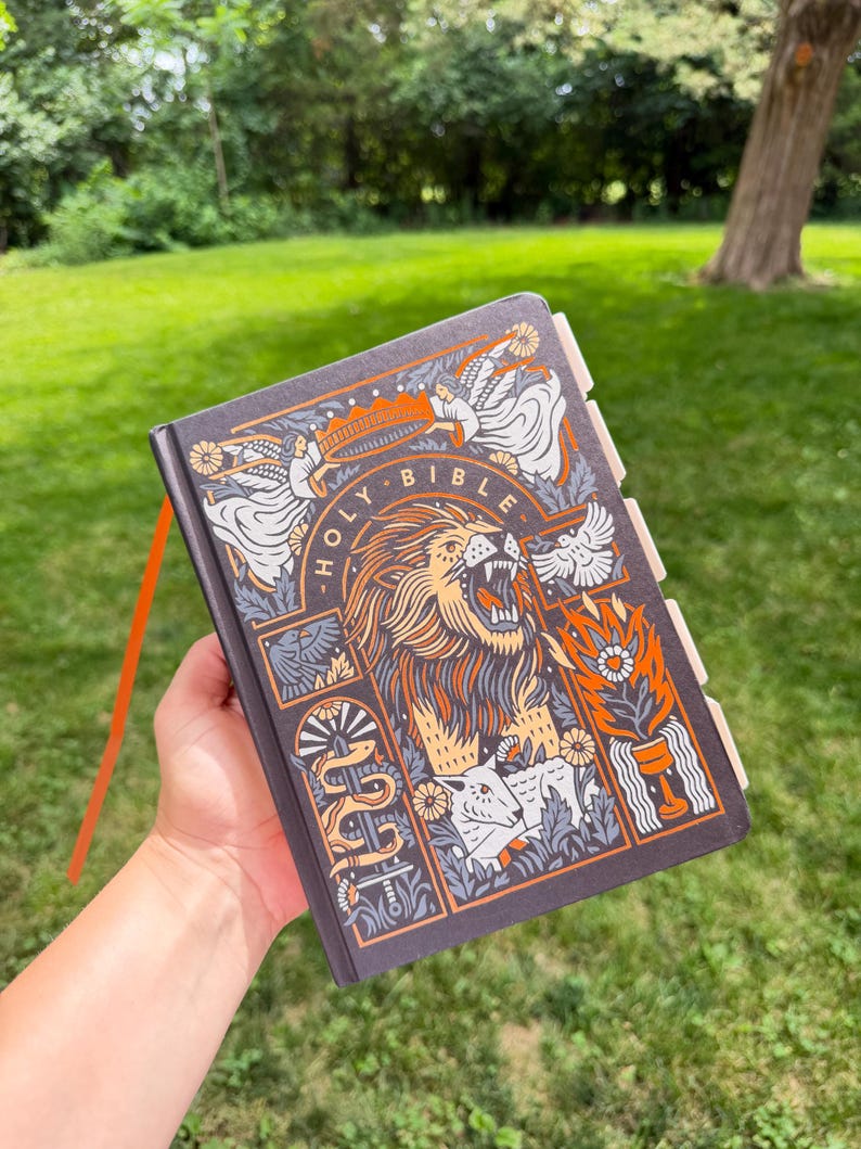 May include: A dark gray hardcover Holy Bible featuring a detailed graphic illustration of a roaring lion, a lamb, angels, and a sword. The cover design uses orange, white, and gray ink. The book includes a bright orange ribbon bookmark and tabs.