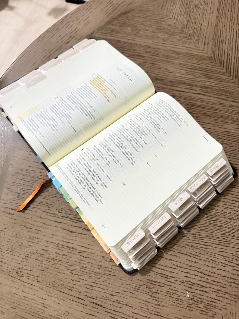 May include: An open Bible with cream-colored pages, featuring printed text, lined margins for notes, and a series of labeled index tabs along the side. The tabs include visible text such as Proverbs, Jeremiah, Isaiah, Song of Solomon, and Ecclesiastes.