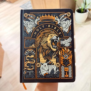 May include: A black Holy Bible with a detailed orange and white graphic illustration on the cover. The design features a roaring lion, a crown, angels, a lamb, a snake, a bird, and flames. The book has visible page tabs along the right side.