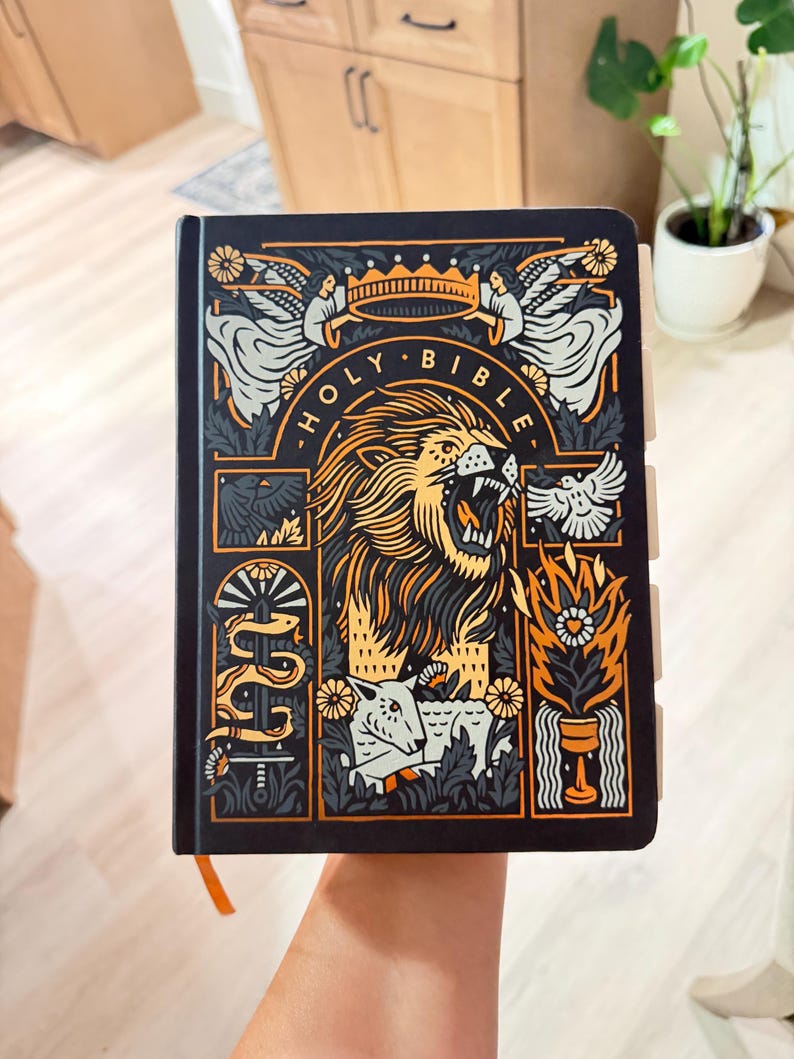 May include: A black Holy Bible with a detailed orange and white graphic illustration on the cover. The design features a roaring lion, a crown, angels, a lamb, a snake, a bird, and flames. The book has visible page tabs along the right side.