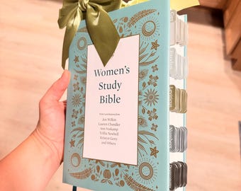 May include: A light blue study Bible with gold botanical illustrations on the cover. The text reads Women's Study Bible with contributions from Jen Wilkin, Lauren Chandler, Ann Voskamp, Trillia Newbell, Kristyn Getty and others. It features a green ribbon bow.