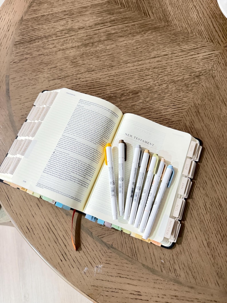 May include: An open Bible with the text New Testament visible on the page. The book features numerous light-colored index tabs along the edges. Six thin markers in shades of yellow, brown, gray, beige, peach, and light blue rest on the open pages.