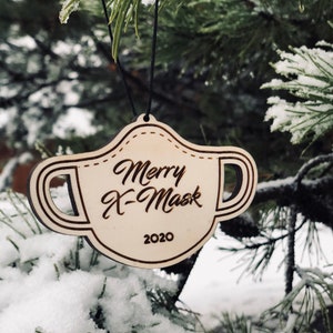 May include: A wooden Christmas ornament shaped like a face mask, hanging from a black string. The ornament has the words "Merry X-Mask 2020" engraved on it. The ornament is hanging on a snow-covered pine tree.