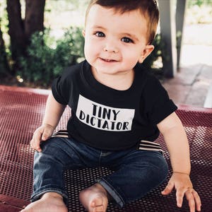 以下が含まれることがあります： 白い長方形の中に「TINY DICTATOR」という文字が入った黒いTシャツを着た笑顔の赤ちゃん。赤ちゃんは青いジーンズも履いており、赤い金属の表面に座っています。