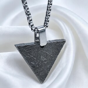 May include: A triangular pendant with a dark, textured, metallic surface featuring a complex, etched pattern. The pendant is attached to a silver-toned box chain necklace by a smooth, polished metal bail, creating a contrast in texture.