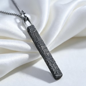May include: A dark grey, rectangular bar pendant with a deeply textured, rugged surface. The pendant hangs from a matching dark grey metal chain, featuring a simple, polished metal bail that connects the bar to the chain link structure.
