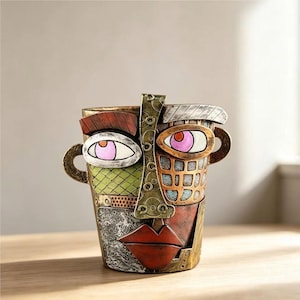 Può includere: Un vaso decorativo a forma di volto umano con finitura multicolore. Il volto presenta un naso centrale dorato, labbra rosse e due occhi con iridi rosa. La superficie mostra varie texture, tra cui tratteggi verdi e motivi metallici.