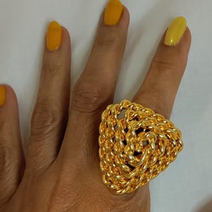 Peut inclure: Une grosse bague dor&eacute;e avec un motif de corde tress&eacute;e en forme triangulaire. La bague est port&eacute;e sur une main avec du vernis &agrave; ongles jaune. La bague est un bijou de caract&egrave;re.
