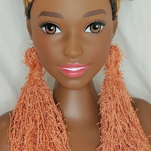 May include: A mannequin head with brown skin, brown eyes, and a full head of dark brown curly hair with blonde highlights. The mannequin is wearing large, orange fringe earrings. The lips are painted pink.