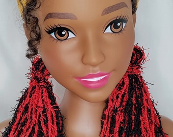 Puede incluir: Una cabeza de muñeca con piel marrón oscura, ojos marrones y labios rosa brillante. El cabello está peinado en un moño alto y voluminoso con rizos en tonos marrón oscuro y rubio dorado. La muñeca lleva pendientes grandes de flecos rojos.