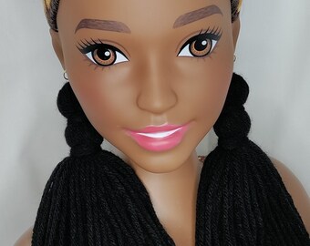 Puede incluir: Cabeza de maniquí con piel morena, ojos marrones y labios rosados. El cabello es una mezcla de marrón, negro y rubio, peinado en un moño alto con dos trenzas de hilo negro. Se ven pequeños pendientes de aro.