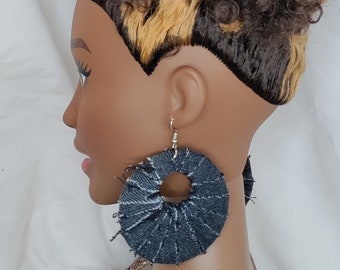 Op de afbeelding: Een mannequinhoofd met donkerbruin krullend haar, geaccentueerd met blonde highlights. De mannequin draagt grote, cirkelvormige denim oorbellen met een gerafelde rand. De oorbellen hebben een zilveren haaksluiting.