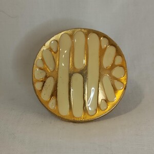 K&ouml;nnte beinhalten: Runde, goldfarbene Brosche mit cremefarbenem Emaille-Design. Die Brosche hat ein zentrales vertikales Design, das von einer Reihe cremefarbener, bl&uuml;tenblatt&auml;hnlicher Formen flankiert wird. Vintage-Accessoire.