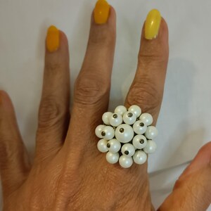 May include: A cluster ring with multiple small, round, white beads. The ring is on a hand with yellow nail polish. The beads are arranged closely, forming a floral or clustered pattern. The ring is a statement piece.