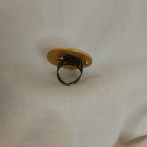 K&ouml;nnte beinhalten: Ein goldfarbener Ring mit einer gro&szlig;en, strukturierten, ovalen Oberfl&auml;che. Das Band des Rings ist aus dunklem, verziertem Metall, und die Oberfl&auml;che hat ein geh&auml;mmertes Aussehen. Der Ring ist vor einem wei&szlig;en Hintergrund platziert.