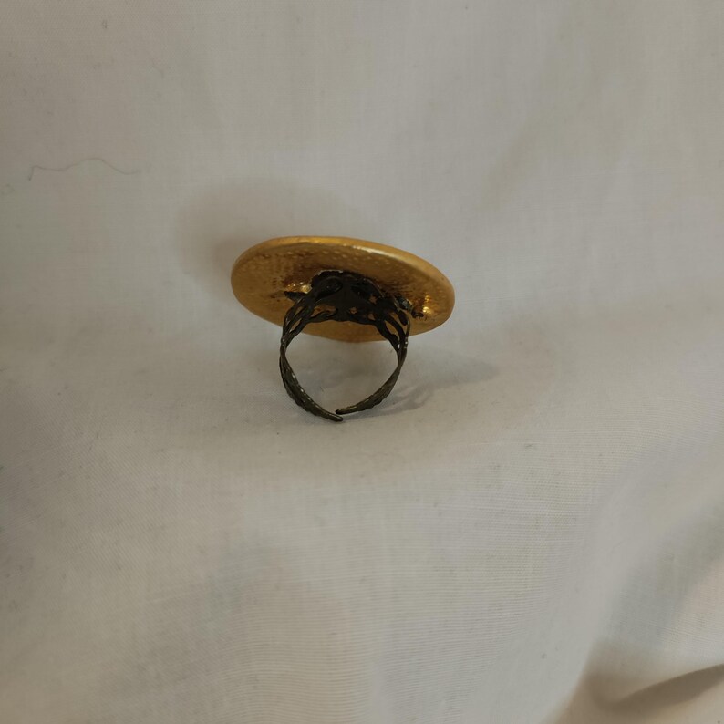 K&ouml;nnte beinhalten: Ein goldfarbener Ring mit einer gro&szlig;en, strukturierten, ovalen Oberfl&auml;che. Das Band des Rings ist aus dunklem, verziertem Metall, und die Oberfl&auml;che hat ein geh&auml;mmertes Aussehen. Der Ring ist vor einem wei&szlig;en Hintergrund platziert.