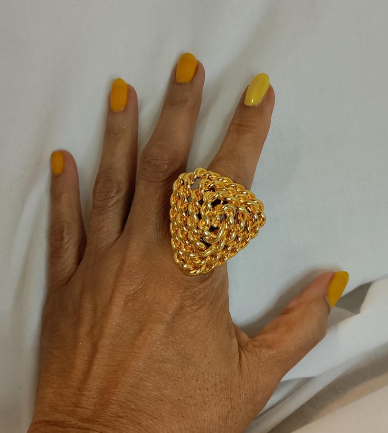 Peut inclure: Une grosse bague dor&eacute;e avec un motif de corde tress&eacute;e en forme triangulaire. La bague est port&eacute;e sur une main avec du vernis &agrave; ongles jaune. La bague est un bijou de caract&egrave;re.