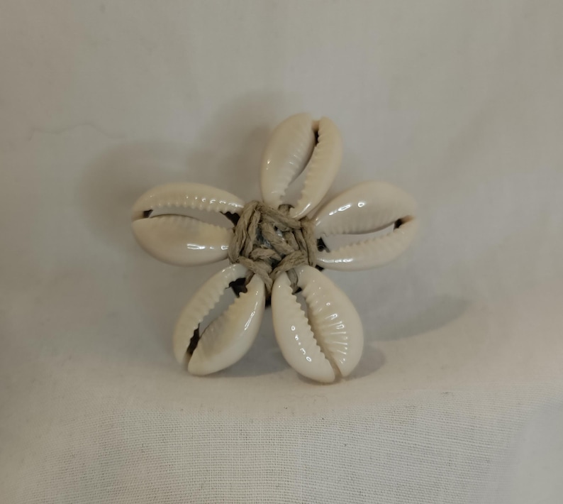 May include: A flower-shaped ring made from five white seashells, each with a natural, glossy finish. The shells are arranged around a central knot of beige twine, creating a unique, beach-inspired accessory. The ring is set against a plain white background.