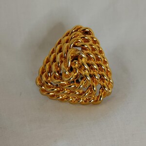 Peut inclure: Broche triangulaire dor&eacute;e avec un motif de corde tress&eacute;e. La broche est en m&eacute;tal brillant et pr&eacute;sente une texture en trois dimensions. Le bijou est pos&eacute; sur un fond blanc.