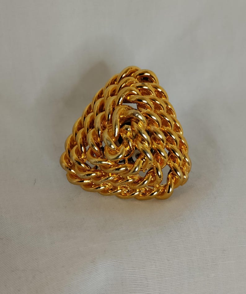 Peut inclure: Broche triangulaire dor&eacute;e avec un motif de corde tress&eacute;e. La broche est en m&eacute;tal brillant et pr&eacute;sente une texture en trois dimensions. Le bijou est pos&eacute; sur un fond blanc.