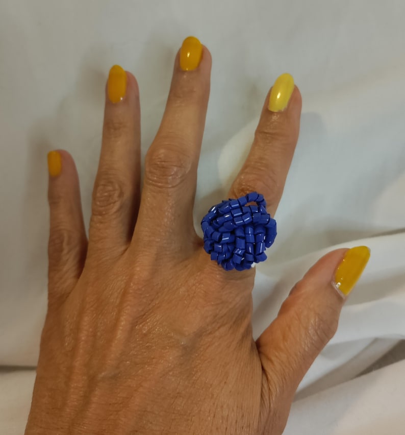 May include: A handcrafted ring featuring a woven design of small, vibrant blue beads. The ring is a bold statement piece, showcasing a unique texture and color. The nails are painted with yellow nail polish.