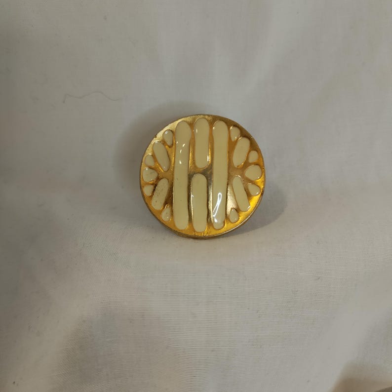 K&ouml;nnte beinhalten: Runde, goldfarbene Brosche mit cremefarbenem Emaille-Design. Die Brosche hat ein zentrales vertikales Design, das von einer Reihe cremefarbener, bl&uuml;tenblatt&auml;hnlicher Formen flankiert wird. Vintage-Accessoire.