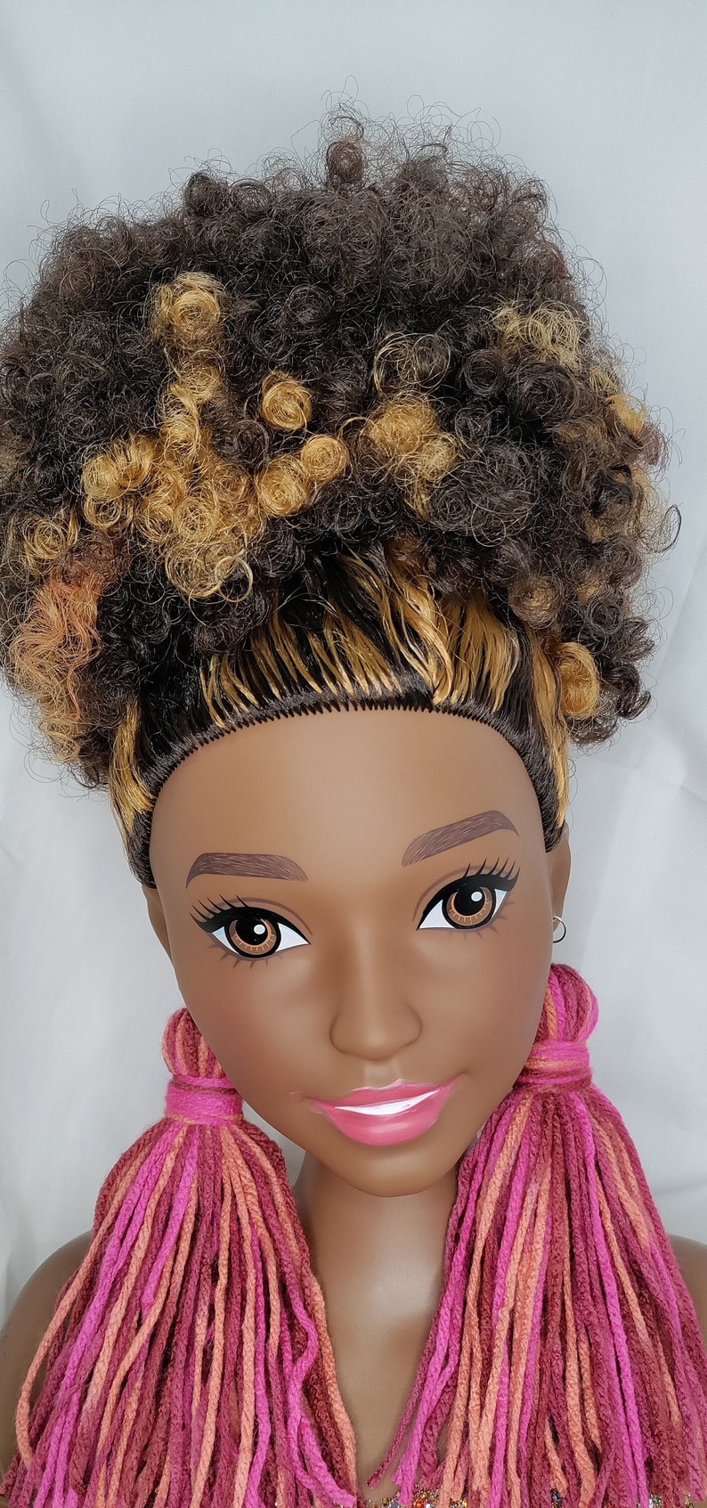 May include: A doll with brown skin and brown and blonde curly hair wearing long pink, orange, and red yarn tassel earrings.
