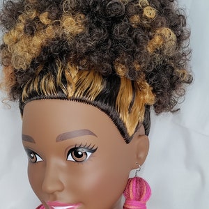 May include: A doll with brown and blonde curly hair wearing long pink, orange, and brown yarn tassel earrings.