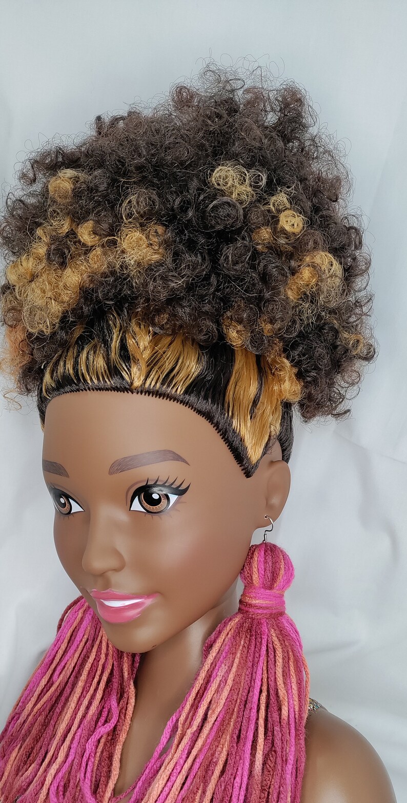 May include: A doll with brown and blonde curly hair wearing long pink, orange, and brown yarn tassel earrings.