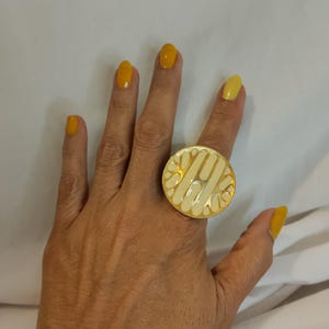 K&ouml;nnte beinhalten: Ein goldfarbener Ring mit einer gro&szlig;en, kreisf&ouml;rmigen Oberfl&auml;che. Die Ringoberfl&auml;che hat ein cremefarbenes Emaille-Design mit einem geometrischen Muster. Der Ring wird an einem Finger mit gelbem Nagellack getragen.