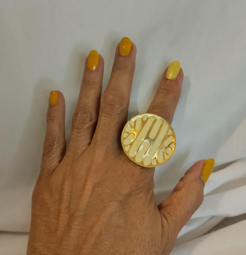 K&ouml;nnte beinhalten: Ein goldfarbener Ring mit einer gro&szlig;en, kreisf&ouml;rmigen Oberfl&auml;che. Die Ringoberfl&auml;che hat ein cremefarbenes Emaille-Design mit einem geometrischen Muster. Der Ring wird an einem Finger mit gelbem Nagellack getragen.