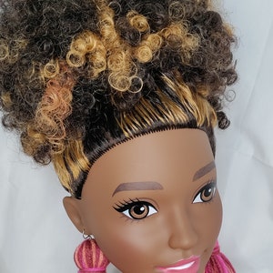 May include: A doll with brown and blonde curly hair wearing long pink, orange, and red yarn tassel earrings.