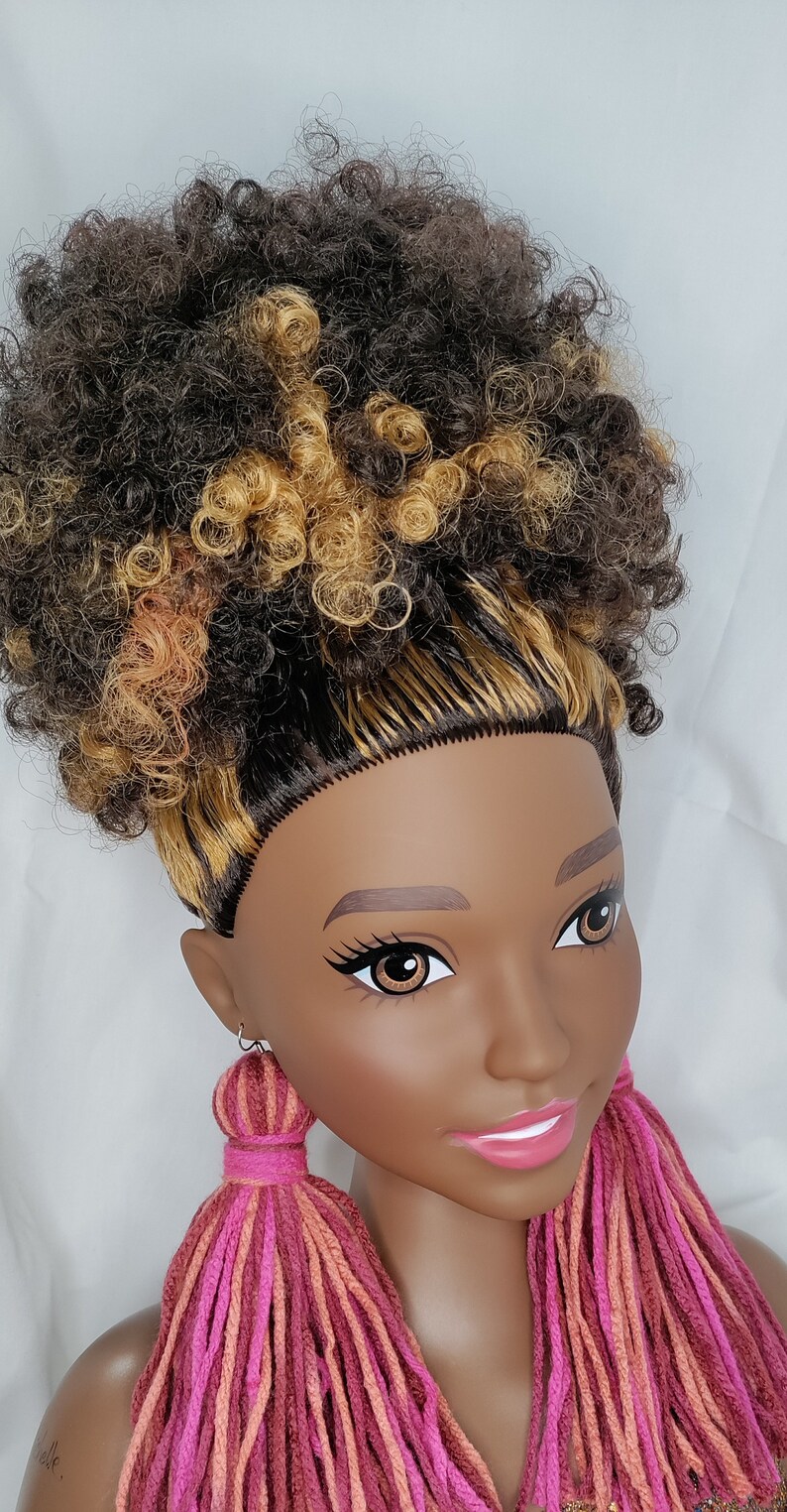 May include: A doll with brown and blonde curly hair wearing long pink, orange, and red yarn tassel earrings.