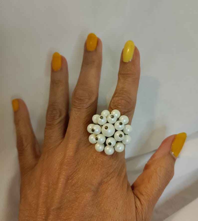 May include: A cluster ring with multiple small, round, white beads. The ring is on a hand with yellow nail polish. The beads are arranged closely, forming a floral or clustered pattern. The ring is a statement piece.