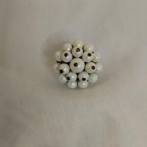May include: A vintage brooch composed of numerous off-white, spherical beads. Each bead has a dark central detail, contrasting with the lighter hue. The brooch is likely made of plastic or glass beads, and is a classic accessory.