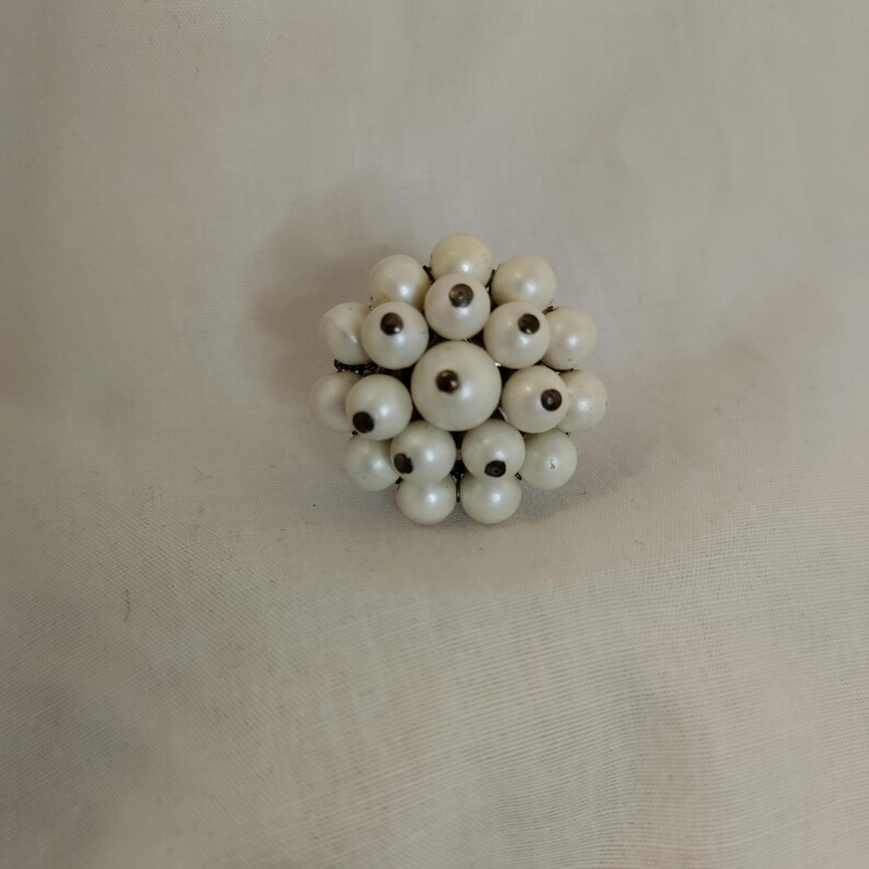 May include: A vintage brooch composed of numerous off-white, spherical beads. Each bead has a dark central detail, contrasting with the lighter hue. The brooch is likely made of plastic or glass beads, and is a classic accessory.