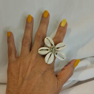 May include: A large, flower-shaped ring made of white seashells. The ring's petals are formed by the shells, and the center is bound with a natural-colored material. The hand wearing the ring has yellow nail polish.