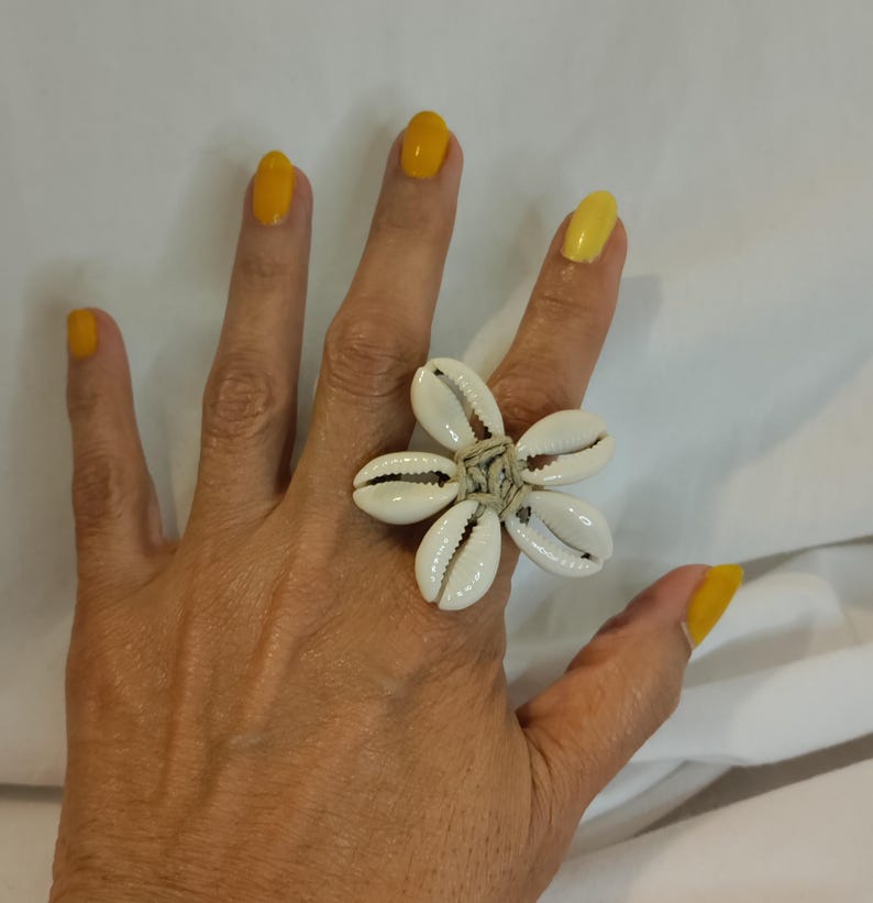 May include: A large, flower-shaped ring made of white seashells. The ring's petals are formed by the shells, and the center is bound with a natural-colored material. The hand wearing the ring has yellow nail polish.