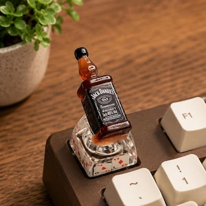Peut inclure: Une touche de clavier mécanique personnalisée présentant une bouteille miniature de whisky Jack Daniel's Tennessee fixée sur une base transparente ornée d'un motif de cartes à jouer rouges et noires, incluant cœurs, carreaux, trèfles et piques.