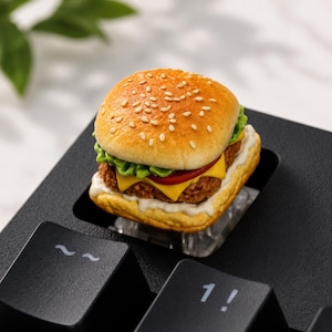 Puede incluir: Una tecla de teclado en miniatura con forma de hamburguesa que presenta un pan dorado con semillas de sésamo, lechuga verde, una rodaja de tomate rojo, una loncha de queso amarillo y una carne marrón sobre una base de salsa blanca.